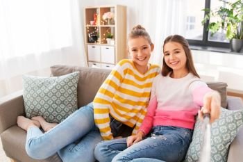 people, technology and friendship concept - happy teenage girls taking picture with selfie stick sitting on sofa at home. girls taking picture with selfie stick at home