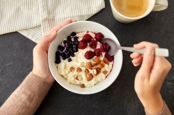 food and breakfast concept - hands of woman with spoon eating porridge in bowl with wild berries, almond nuts and cup of coffee on slate stone table. hands with porridge breakfast and cup of coffee