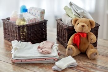 babyhood and clothing concept - baby clothes, teddy bear toy and wicker baskets on wooden table at home. baby clothes and teddy bear toy on table at home