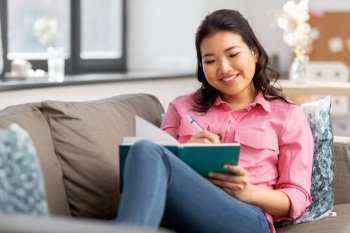 people and leisure concept - happy smiling asian young woman with diary or notebook sitting on sofa at home. asian woman with diary sitting on sofa at home