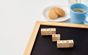 quarantine, epidemic and safety concept - close up of chalkboard with stay at home words on wooden toy blocks, coffee cup, cookies and aroma reed diffuser on white background. chalkboard with stay at home words on toy blocks