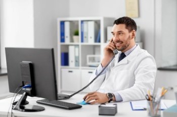 healthcare, medicine and people concept - smiling male doctor with clipboard calling on desk phone at hospital. male doctor calling on desk phone at hospital