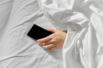 technology, comfort and morning concept - hand of young woman lying in bed with smartphone at bedroom. hand of woman lying in bed with smartphone