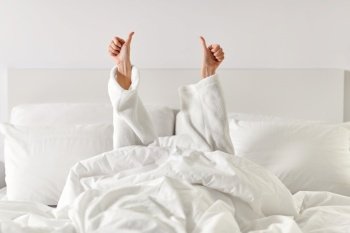 gesture, comfort and morning concept - hands of young woman in hotel robe lying in bed and showing thumbs up at bedroom. hands of woman lying in bed and showing thumbs up