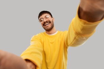 people concept - happy smiling young man in glasses and yellow sweatshirt making selfie over grey background. happy young man in yellow sweatshirt making selfie