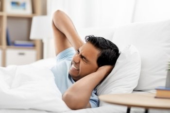 people, bedtime and rest concept - annoyed indian man lying in bed at home and covering ears. annoyed indian man lying in bed and closing ears