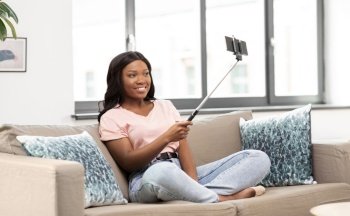 technology and people concept - happy smiling african american woman with smartphone and selfie stick taking picture sitting on sofa at home. happy african american woman taking selfie at home
