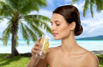 beauty and detox concept - woman drinking fresh water with lemon and ice over tropical beach background in french polynesia. woman drinking ice water with lemon over beach