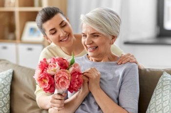 family, mother’s day and birthday concept - happy smiling adult daughter giving flowers to her senior mother at home. adult daughter giving flowers to old mother