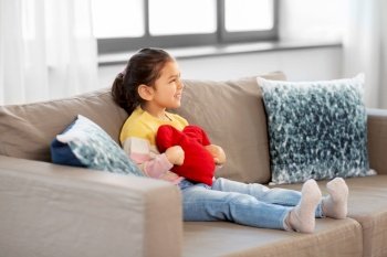 childhood, valentine’s day and people concept - happy little girl with heart shaped pillow sitting on sofa at home. happy little girl with heart shaped pillow at home