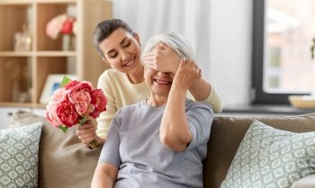family, mother’s day and birthday concept - happy smiling adult daughter giving flowers to her senior mother at home. adult daughter giving flowers to old mother