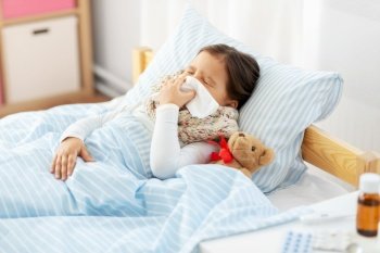 health, children and people concept - sick little girl lying in bed and blowing nose with paper tissue at home. sick girl lying in bed and blowing nose at home