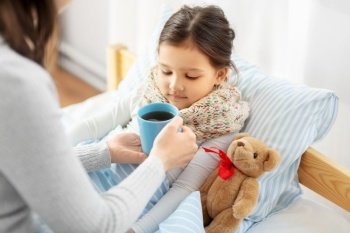 family, health and people concept - mother giving mug with hot tea to sick little daughter lying in bed at home. mother giving hot tea to sick little daughter