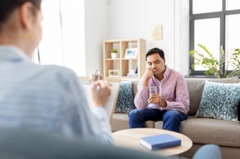 psychology, mental therapy and people concept - sad young indian man patient with glass of water and woman psychologist at psychotherapy session. man and psychologist at psychotherapy session