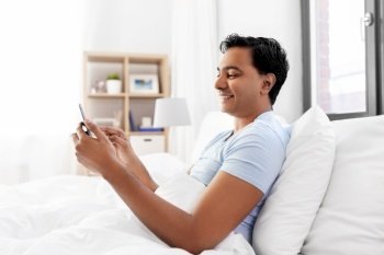 technology, bedtime and rest concept - happy smiling indian man with smartphone lying in bed at home. happy indian man with smartphone in bed at home