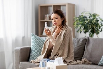 health, cold and people concept - sad sick young asian woman in blanket with oral spray medicine at home. sick asian woman with oral spray medicine at home