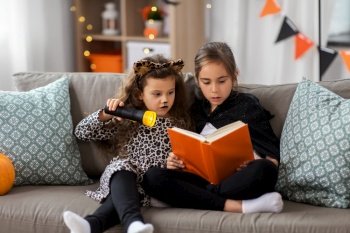 halloween, holiday and childhood concept - little girls in costumes with flashlight reading book at home. girls in halloween costumes reading book at home