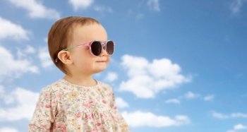 childhood, summer and people concept - happy little baby girl in sunglasses over blue sky and clouds background. happy little baby girl in sunglasses over blue sky