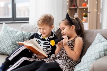 halloween, holiday and childhood concept - little kids in party costumes with flashlight reading book at home. kids in halloween costumes reading book at home