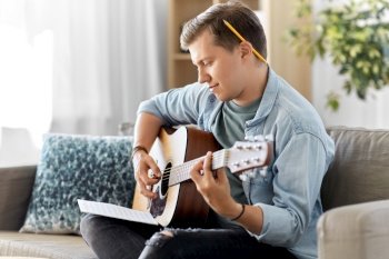 leisure and people concept - young man or musician with music book playing guitar sitting on sofa at home. young man with music book playing guitar at home