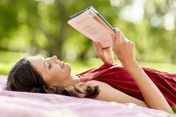 leisure and people concept - happy smiling woman reading book lying on picnic blanket at summer park. happy woman reading book lying on blanket at park