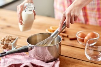 cooking, culinary and seasonal drinks concept - close up of hand with whisk and milk making eggnog in pot. hands with whisk and milk cooking eggnog