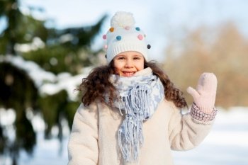 childhood, leisure and season concept - portrait of happy little girl in winter clothes waving hand at park. happy little girl waving hand outdoors in winter