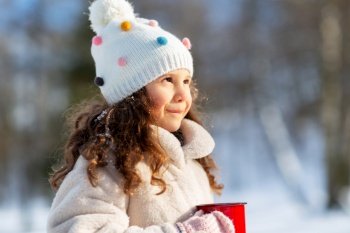 childhood, christmas and season concept - happy little girl with cup of hot tea in winter park. little girl with cup of hot tea in winter park