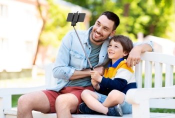 family, fatherhood and people concept - happy father with little son taking picture with smartphone and selfie stick sitting on bench at summer park. father and son taking selfie with phone at park