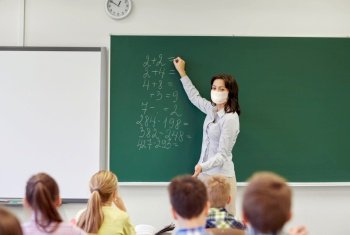 education, school and pandemic concept - female math teacher wearing face protective medical mask for protection from virus disease writing mathematic task on green chalkboard for group of students. teacher in mask writing on chalkboard at school