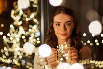 christmas, holidays and people concept - young woman in glasses with festive garland lights in mason jar mug at home at night. woman with christmas garland lights in glass mug