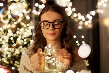 christmas, holiday and people concept - young woman in glasses with festive garland lights in mason jar mug at home. woman with christmas garland lights in glass mug