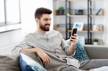 technology, internet, communication and people concept - happy smiling young man texting on smartphone at home. happy smiling young man with smartphone at home