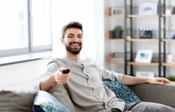 people and leisure concept - happy smiling man with remote control watching tv at home. happy man with remote control watching tv at home