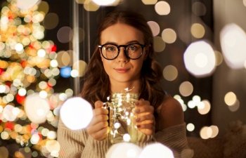 christmas, holidays and people concept - young woman in glasses with garland lights in mason jar mug at home. woman with christmas garland lights in glass mug