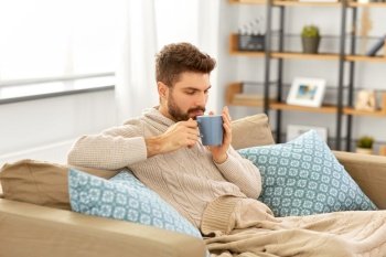 health, cold and people concept - sick young man in blanket drinking hot tea at home. sick young man in blanket drinking hot tea at home