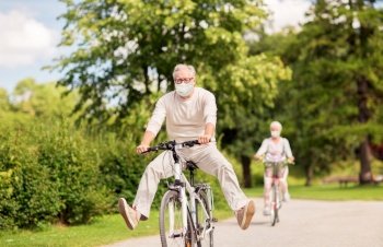old age, health and pandemic concept - happy senior couple wearing face protective medical mask for protection from virus disease riding bicycles at summer park. senior couple in masks riding bicycles at park