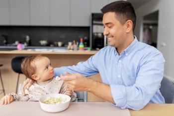 family, food, eating and people concept - happy smiling middle-aged father feeding little baby daughter sitting in highchair with puree by spoon at home. happy father feeding baby in highchair at home