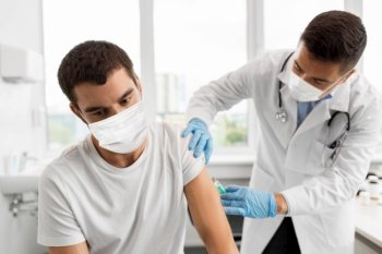 medicine, vaccination and healthcare concept - doctor with syringe doing injection of vaccine to male patient wearing face protective medical masks for protection from virus disease. patient and doctor in masks doing vaccination