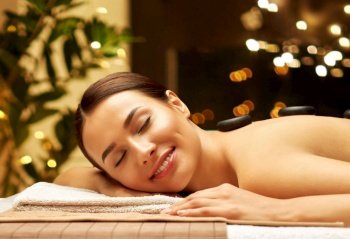 wellness, beauty and relaxation concept - smiling beautiful young woman having hot stone massage at spa over festive lights on background. smiling woman having hot stone massage at spa
