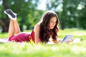 leisure, inspiration and people concept - happy smiling woman writing to diary or notebook lying on picnic blanket at summer park. happy woman with diary or notebook at park