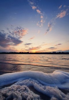 Vertical orientation, golden hour, waterscape. Lot of white fluffy foam on the shore of a pond. Surf created by the lake waves, against colorful evening sky. Idyllic scene, peaceful sunset background.