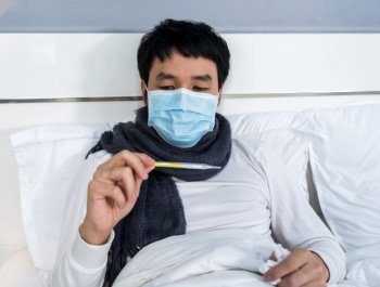 sick man in medical mask using thermometer to checking his temperature on a bed, coronavirus (covid-19) pandemic concept.