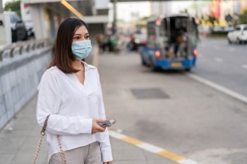 young asian woman holding smartphone and waiting for bus at bus stop in city street and wearing face mask protective for spreading of coronavirus(covid-19) pandemic, new normal concept