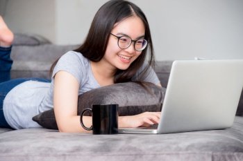 Portrait beautiful Asian young woman lying relax work with a laptop, Cute girl teens looking at the monitor smile happily on the sofa working remotely with an internet computer communication from home. Cute girl relax work with a laptop 