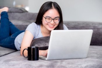 Portrait beautiful Asian young woman lying relax work with a laptop, Cute girl teens looking at the monitor smile happily on the sofa working remotely with an internet computer communication from home. Cute girl relax work with a laptop 