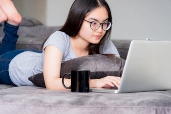 Portrait beautiful Asian young woman lying and relax work with a laptop, Cute girl teens looking at the monitor intently on the sofa working remotely with an internet computer communication from home. Cute girl relax work with a laptop