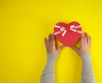 hands  hold a paper closed red box in the shape of a heart on a yellow  background, festive backdrop