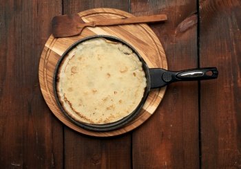 baked round big pancake in a black frying pan on a brown wooden table, top view, carnival