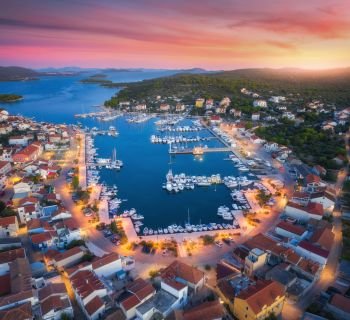 Aerial view of boats and yachts in port and city at sunset. Summer landscape with city lights, buildings, illuminated streets, mountain, motorboats, blue sea, red sky at night. Top view of Croatia